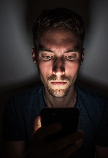 Portrait of a focused man using a smartphone in a dark environment illuminated by screen light.