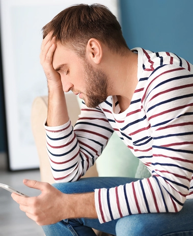 Concerned young man looking at his smartphone while sitting indoors with thoughtful expression.